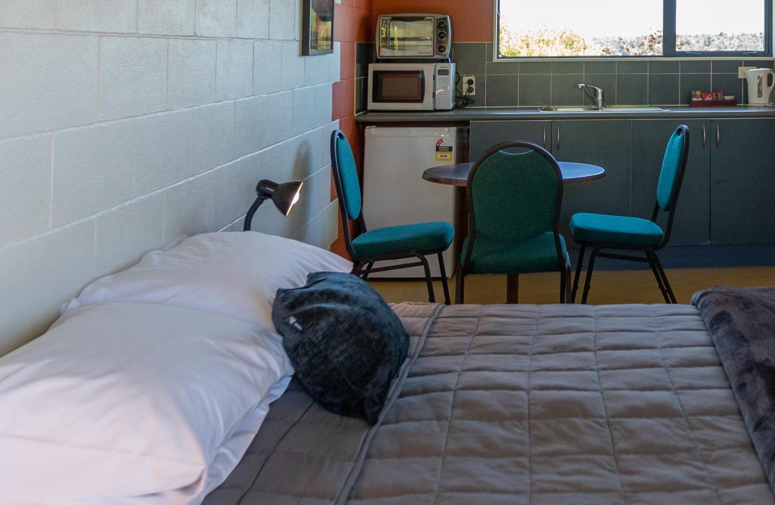 Una cama individual con almohada y manta, una lámpara de noche, una mesita con una silla y una kitchenette con microondas y tostadora.