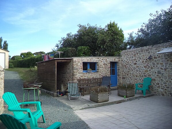 A small stone building with a blue door and window, a wooden structure with a chimney, outdoor seating with chairs, and a gravel path.