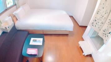 A small, cozy room with a bed, a dark-colored sofa, a small table with a book and a pink item, and a window with curtains.