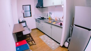 Une cuisine compacte équipée d’un réfrigérateur, d’un évier et d’une cuisinière. On y trouve une fenêtre avec des stores, une petite table avec une chaise et un tapis sur le sol.