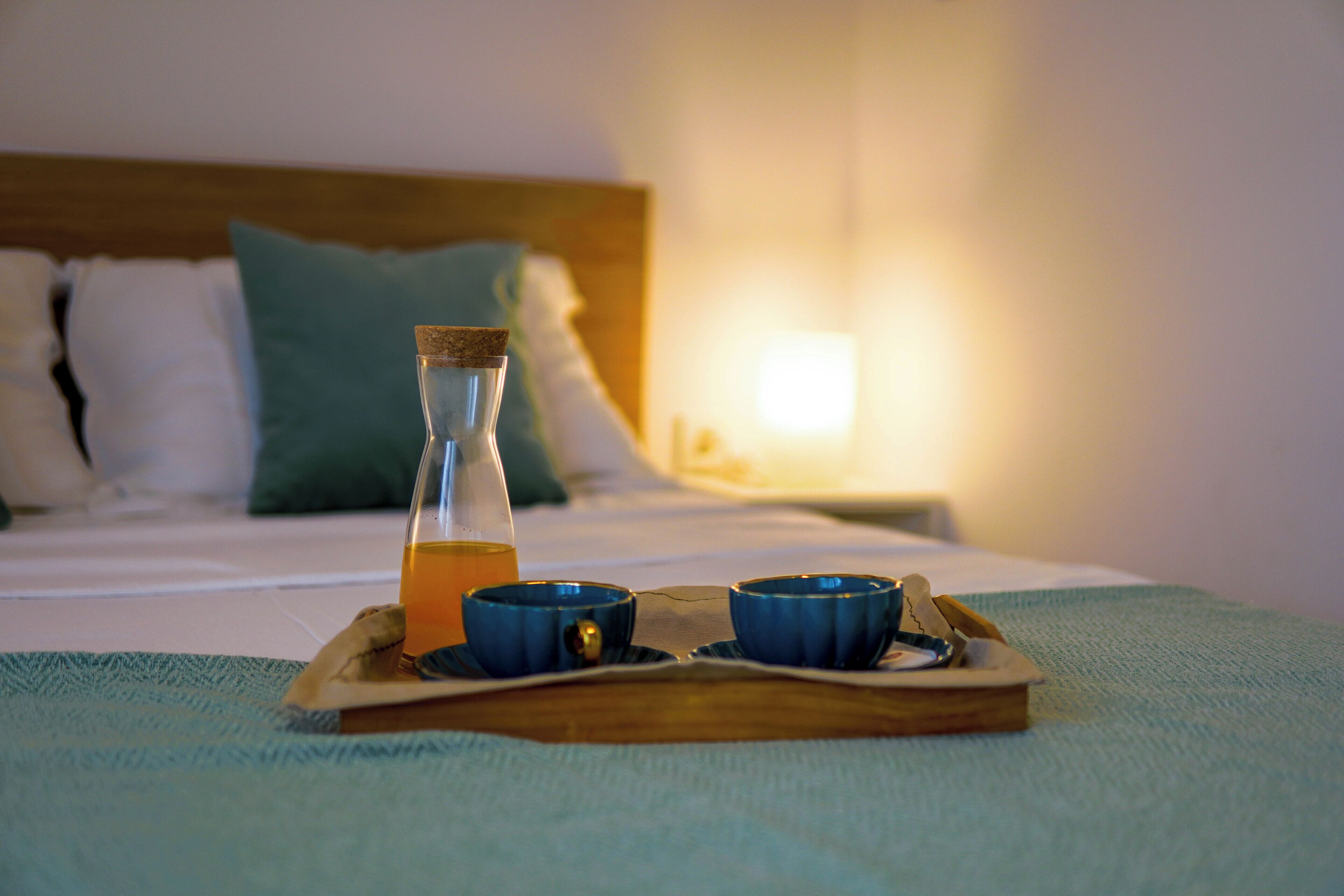 Una bandeja con una botella de vidrio y dos tazas azules sobre una cama con cabecera de madera y almohadas.