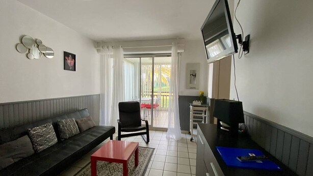 A compact living space with a sofa, a small coffee table, a TV mounted on the wall, and a sliding glass door leading to an outdoor area. - Cozy Ground Floor Studio, Terrace, Parking, Pet-Friendly - Cambo-les-Bains (Cambo-les-Bains)