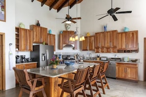 Una cocina con armarios de madera, una isla central con sillas, ventiladores de techo y varios electrodomésticos.
