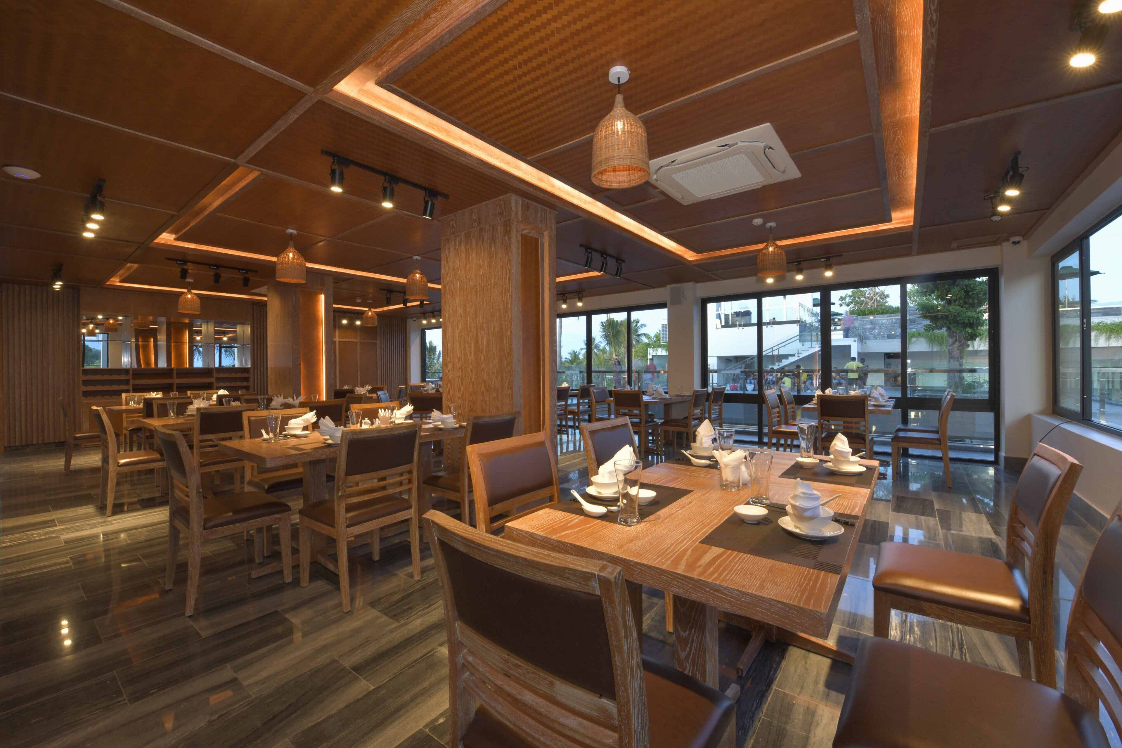 Een ruime eetzaal met houten tafels en stoelen, grote ramen met uitzicht naar buiten en hanglampen.