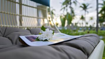 Un libro con una flor blanca sobre un sofá color beige en un entorno exterior, con una valla de bambú y palmeras al fondo.