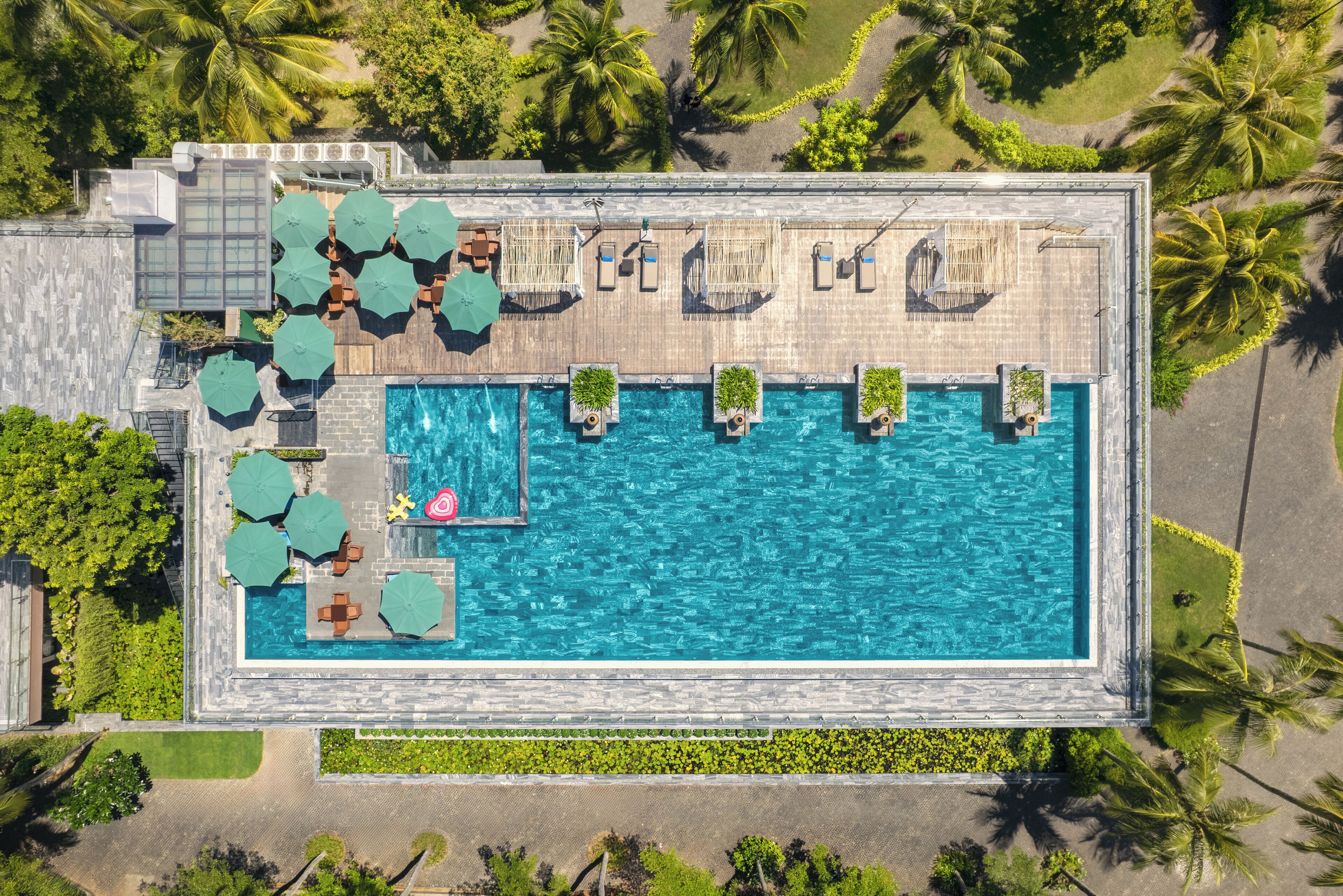 Vue aérienne d’une piscine rectangulaire, agrémentée d’espaces de détente et de parasols, et entourée de palmiers.