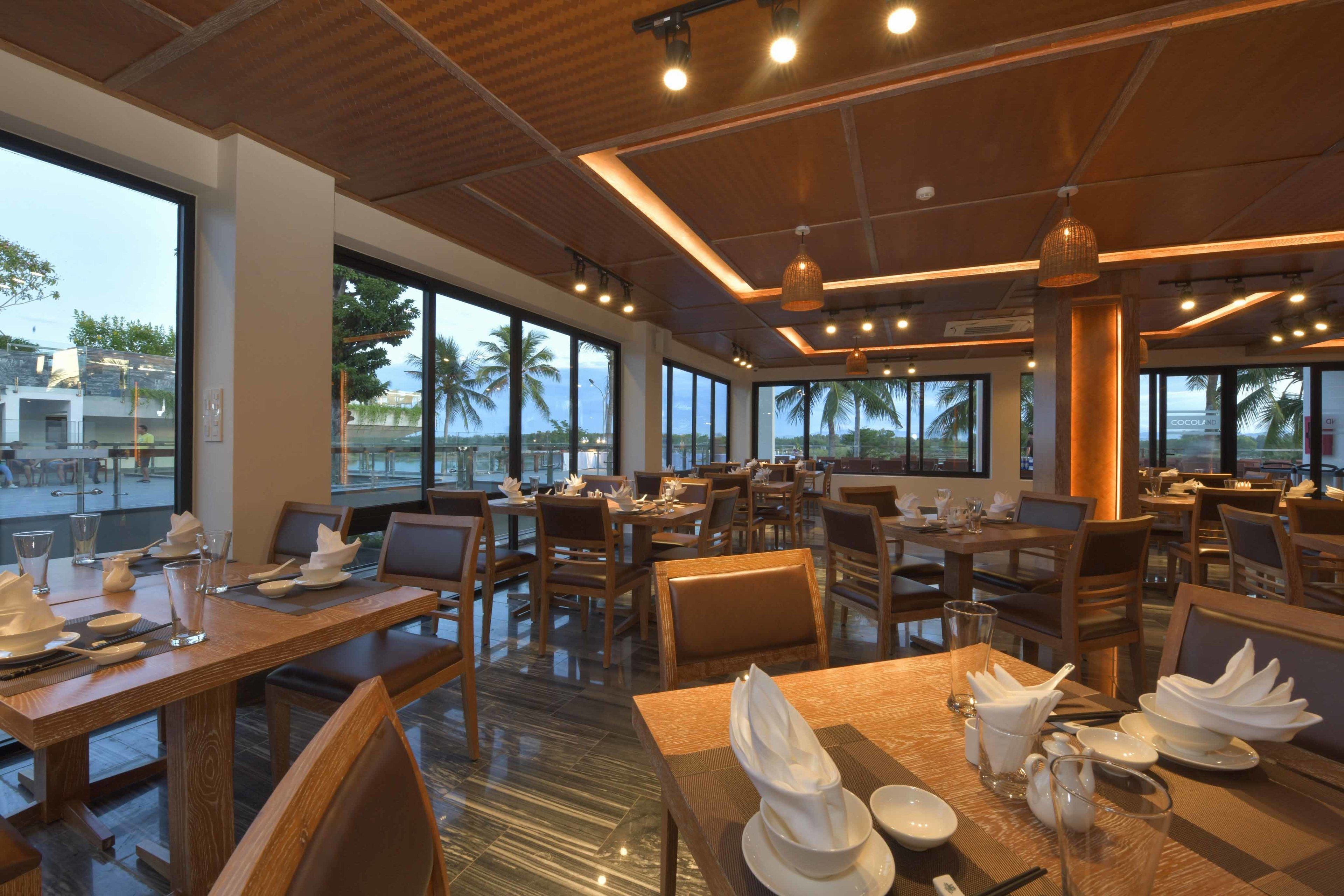 Un restaurante con varias mesas y sillas, ventanas amplias que ofrecen vistas a palmeras y un cuerpo de agua.