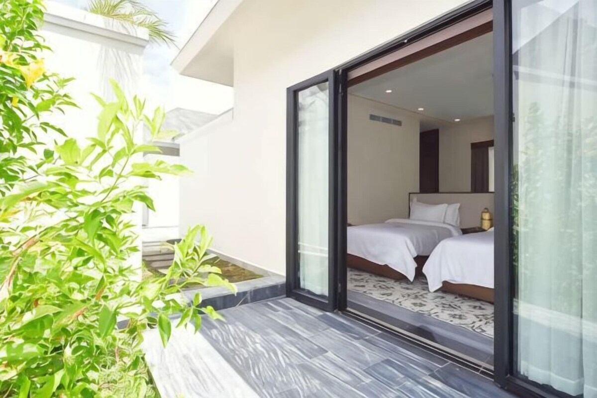 A modern hotel room with a large sliding glass door leading to a balcony, a bed with white bedding, and a small potted plant on the floor.