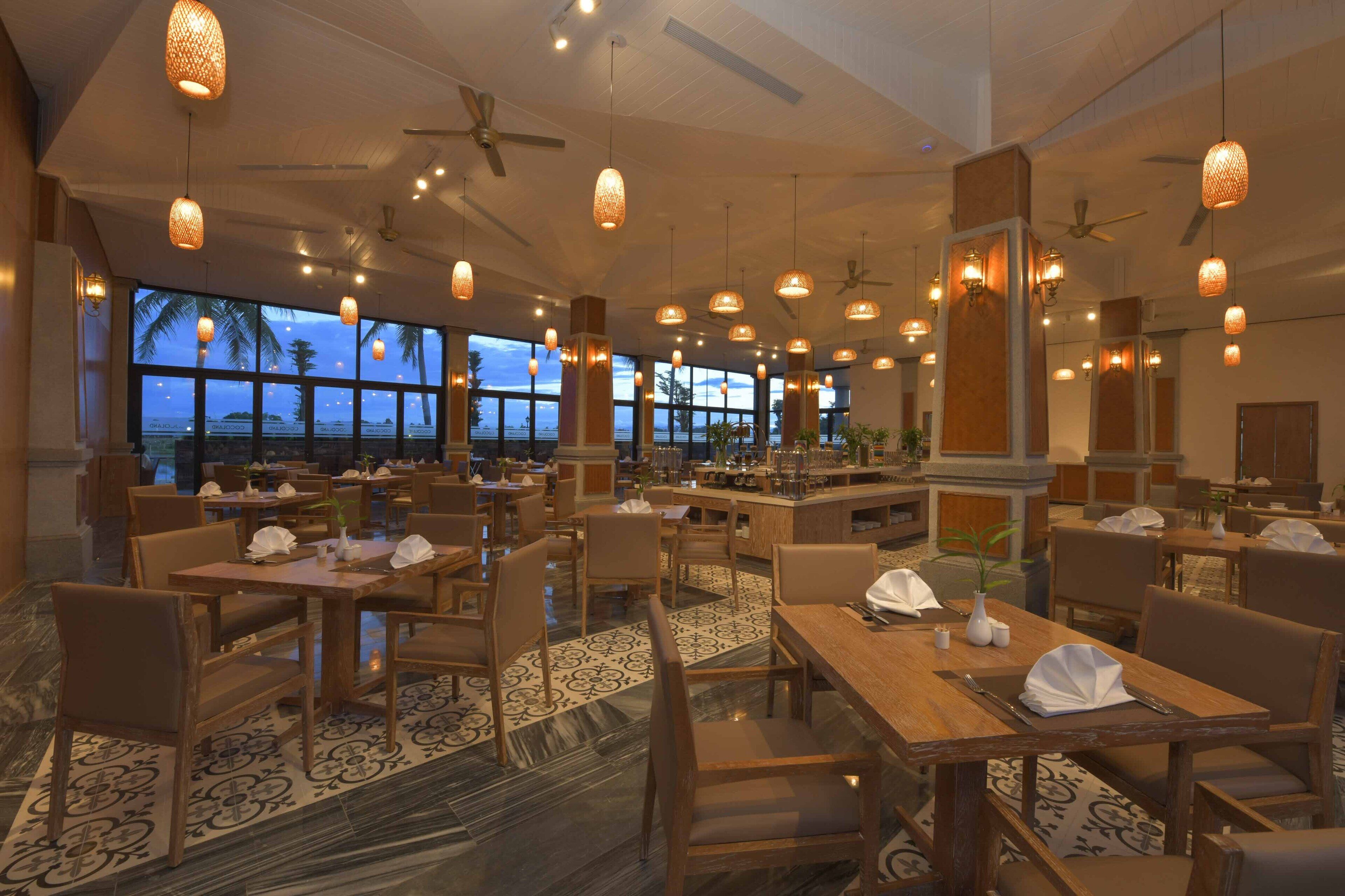 A spacious hotel dining area with multiple tables and chairs, large windows offering a view of the ocean, and hanging pendant lights.