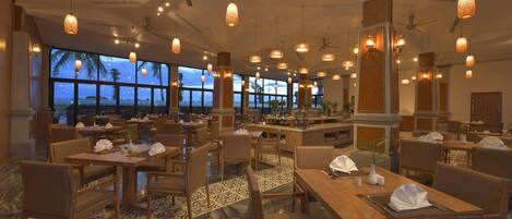 A spacious hotel dining area with multiple tables and chairs, large windows offering a view of the ocean, and hanging pendant lights.