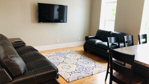 A living room with a black leather sofa, a patterned rug, a flat-screen TV mounted on the wall, and a dining area with a table and chairs. - Mona K's-Little Green Rental (Green Lake)