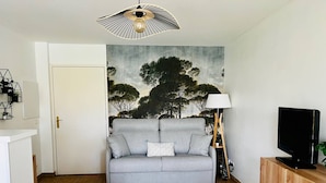 Ein Wohnzimmer mit grauem Sofa, einem Fernseher auf einer Holzkommode, einem Baum-Gemälde an der Wand und einer Lampe neben dem Sofa.