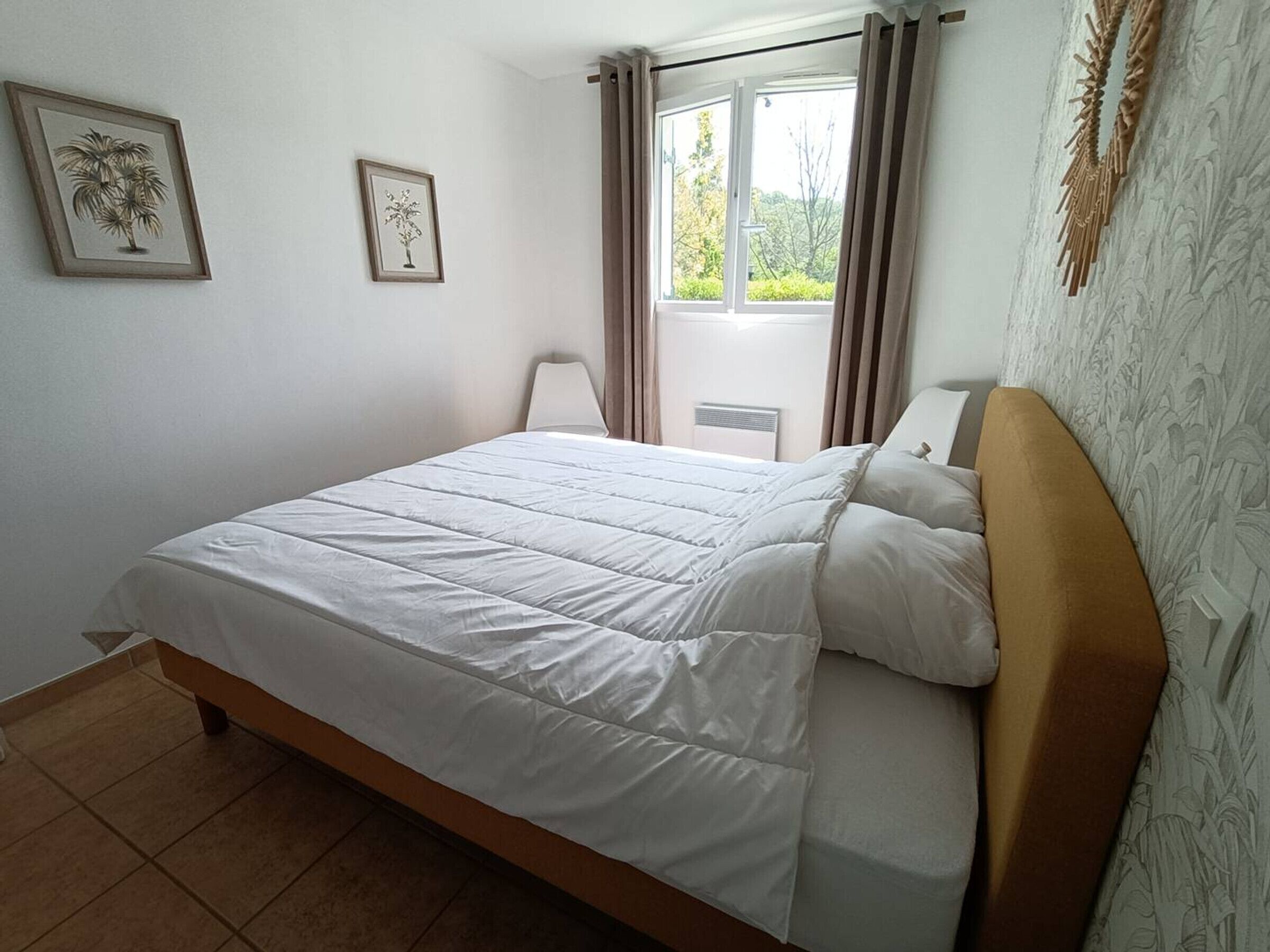 Una cama bien tendida con edredón blanco y almohadas, una mesita de noche con lámpara y dos cuadros enmarcados en la pared.