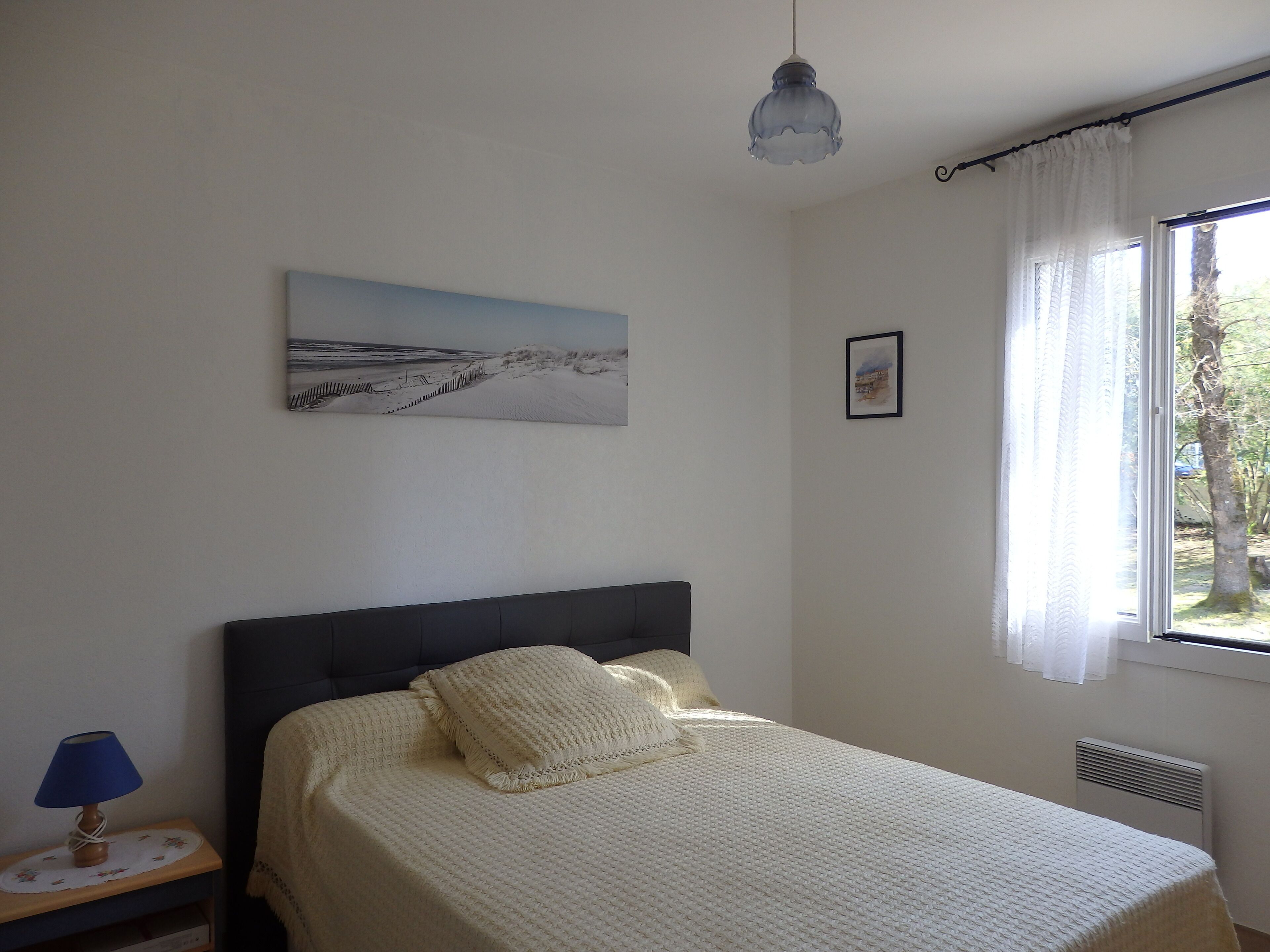 Un dormitorio con una cama, una mesita de noche con lámpara, una ventana con cortinas y un cuadro en la pared.