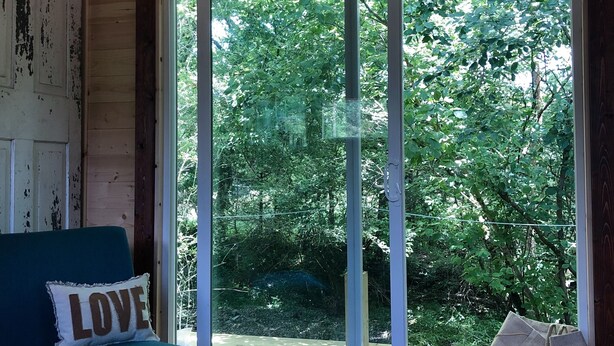 A room with a teal sofa, a "LOVE" pillow, a sliding glass door leading to a wooded area, and a ceiling with a smoke detector.