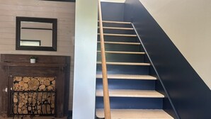 Una scala con gradini in legno e una ringhiera nera, che conduce a un pianerottolo con uno specchio e un camino con legna a vista.