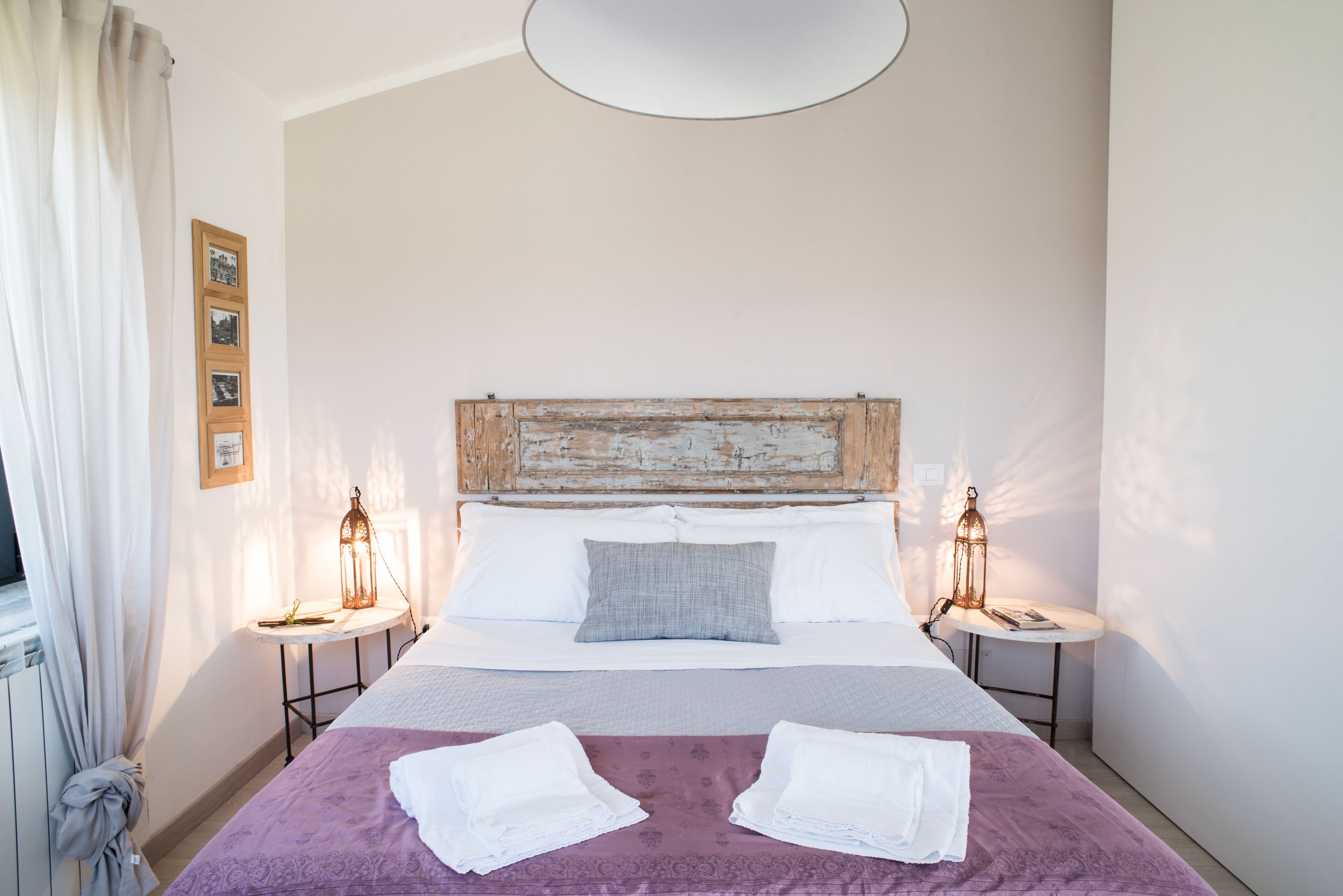Un lit bien fait, recouvert d’une couvre-lit violette, avec deux serviettes blanches et une tête de lit en bois rustique. Deux tables de chevet sont disposées de chaque côté du lit, sur lesquelles reposent des lampes de style lanterne.