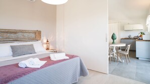 Una camera d'albergo con un letto, un comodino con una lampada, un tavolino con sedie e una piccola cucina in fondo.