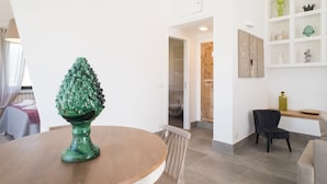 Un'area pranzo con un tavolo rotondo, sedie e un vaso verde. Si nota una sedia in legno e uno scaffale con oggetti decorativi.