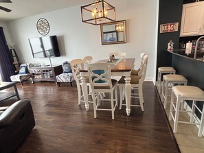 Un coin repas avec une table en bois et des chaises blanches, un téléviseur fixé au mur et un espace cuisine avec des placards.