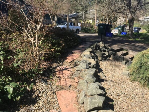 A pathway lined with rocks and a brick path, leading to a parked white truck and a green dumpster.