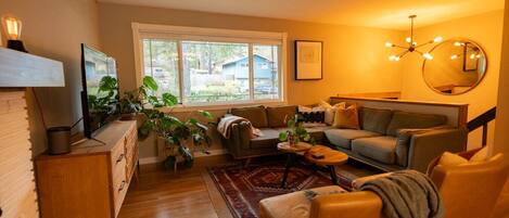 A cozy living room with a sofa, armchairs, a coffee table, and a TV. There is a large window with a view of the outside, and a plant is visible on the windowsill.