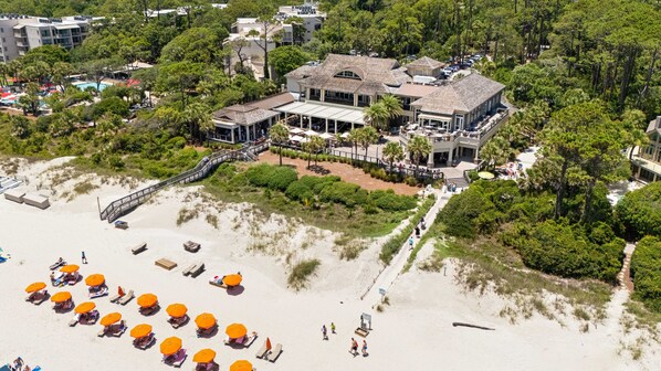 An aerial view of a beachfront property with a large building, a beach with sun umbrellas, and a boardwalk leading to the building. - 21 Shell Ring:  BESTNEST by Beverly Serral Vacation Rentals (Hilton Head Island)