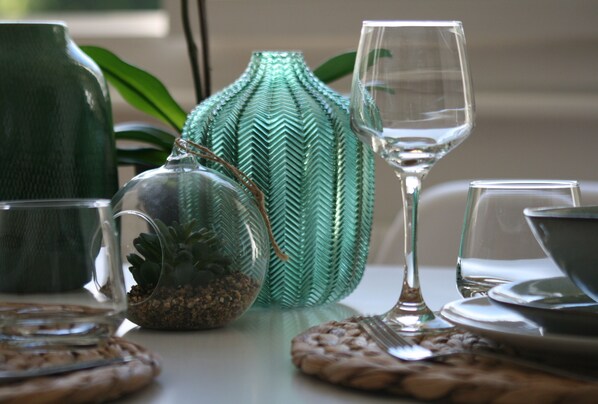 Una mesa de comedor con una jarra de vidrio transparente, un florero de vidrio verde y una copa de vino.