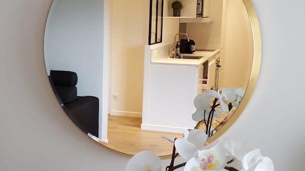 A round mirror reflecting a modern living room with a black sofa, a coffee machine, and a small shelf with decorative items.