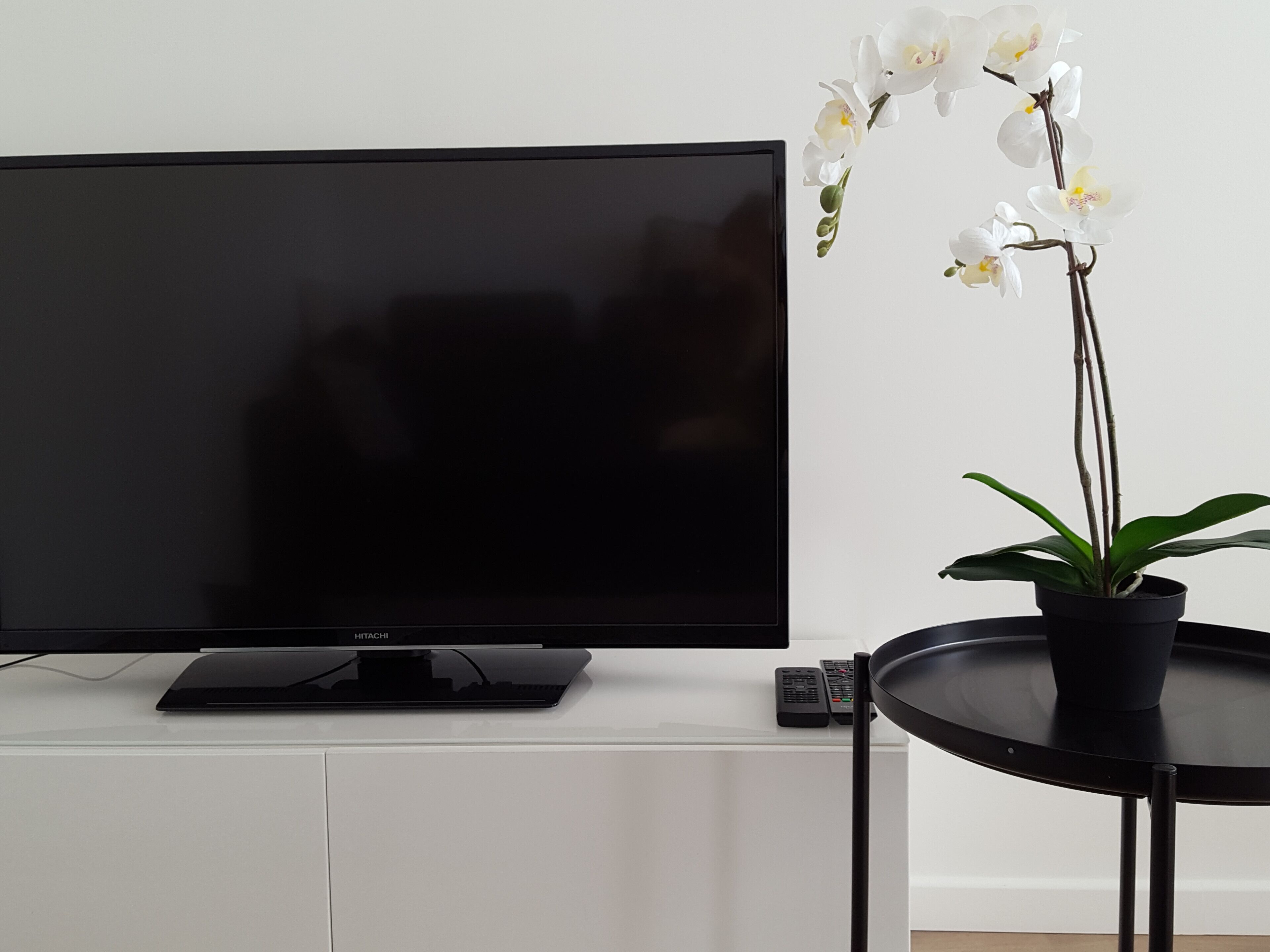 Un téléviseur à écran plat posé sur un meuble blanc, une télécommande et une orchidée en pot sur une petite table.