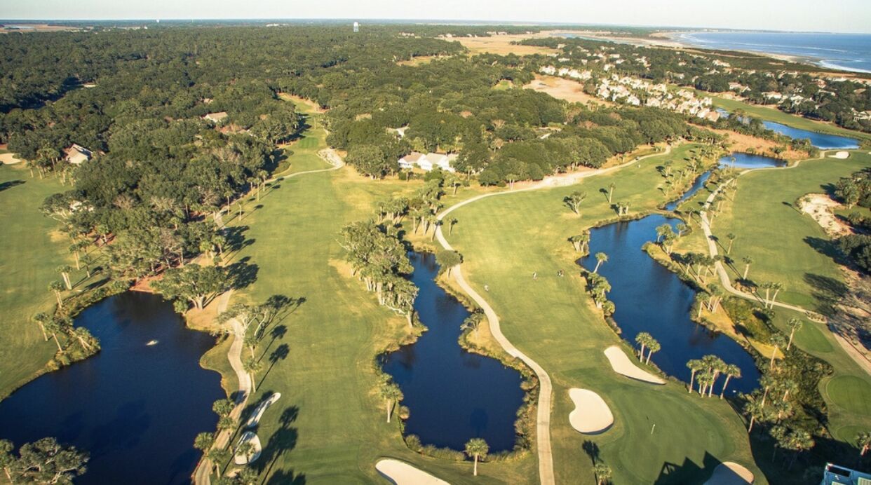 Vista aérea de un campo de golf con varios estanques, bunkers de arena y un bosque denso que lo rodea.