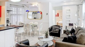 Un salon moderne avec un coin repas, des meubles blancs, un grand miroir et un escalier menant à une autre pièce.