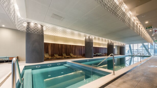 Una piscina interior moderna con baldosas azules cristalinas, una zona de estar con sillones reclinables y vistas a la silueta urbana a través de amplios ventanales.