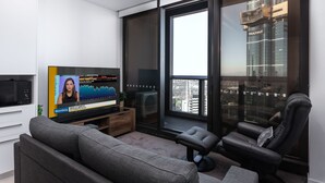 Una sala de estar moderna con un televisor de pantalla plana mostrando un programa de noticias de Bloomberg, un sofá cómodo, un sillón de cuero negro y un ventanal con vistas a la ciudad.