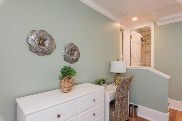 Une salle de bain avec un meuble-lavabo blanc, deux décorations florales murales, une plante en pot, une chaise en osier et un miroir au-dessus du lavabo.