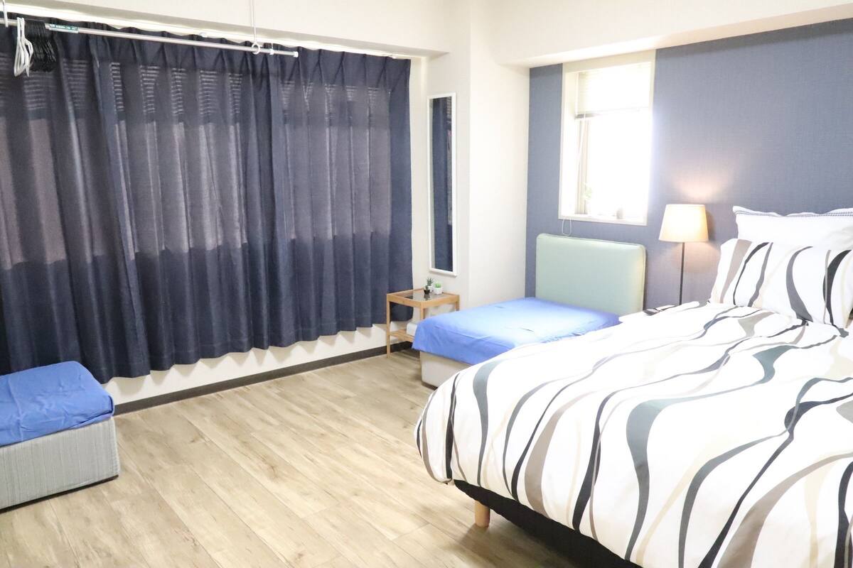 A hotel room with a large bed, a smaller bed, a nightstand, a lamp, a window with curtains, and a wooden floor.