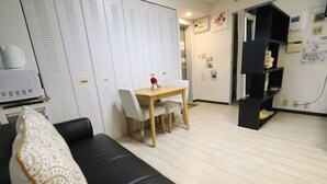A modern living room with a black leather sofa, a wooden dining table with chairs, and a white wardrobe with louvered doors. - Residence Osaka Nipponbashi (Osaka)