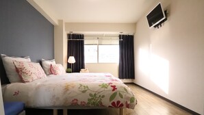 A bedroom with a bed, a window with curtains, a mounted TV, and a lamp. - Residence Osaka Nipponbashi (Osaka)