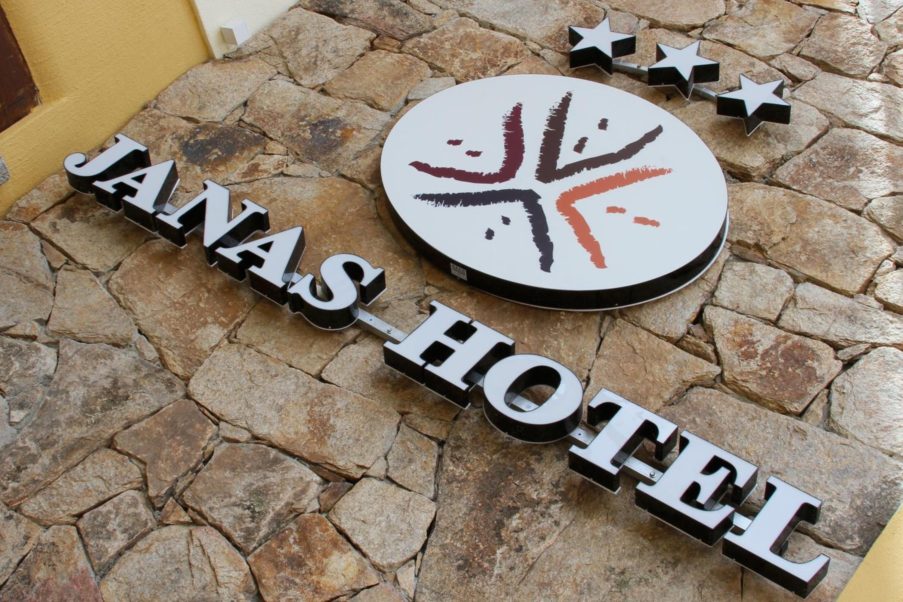 Een stenen muur met een rond bord waarop een gestileerd ontwerp staat afgebeeld en de tekst 'JANAS HOTEL' in vetgedrukte letters.