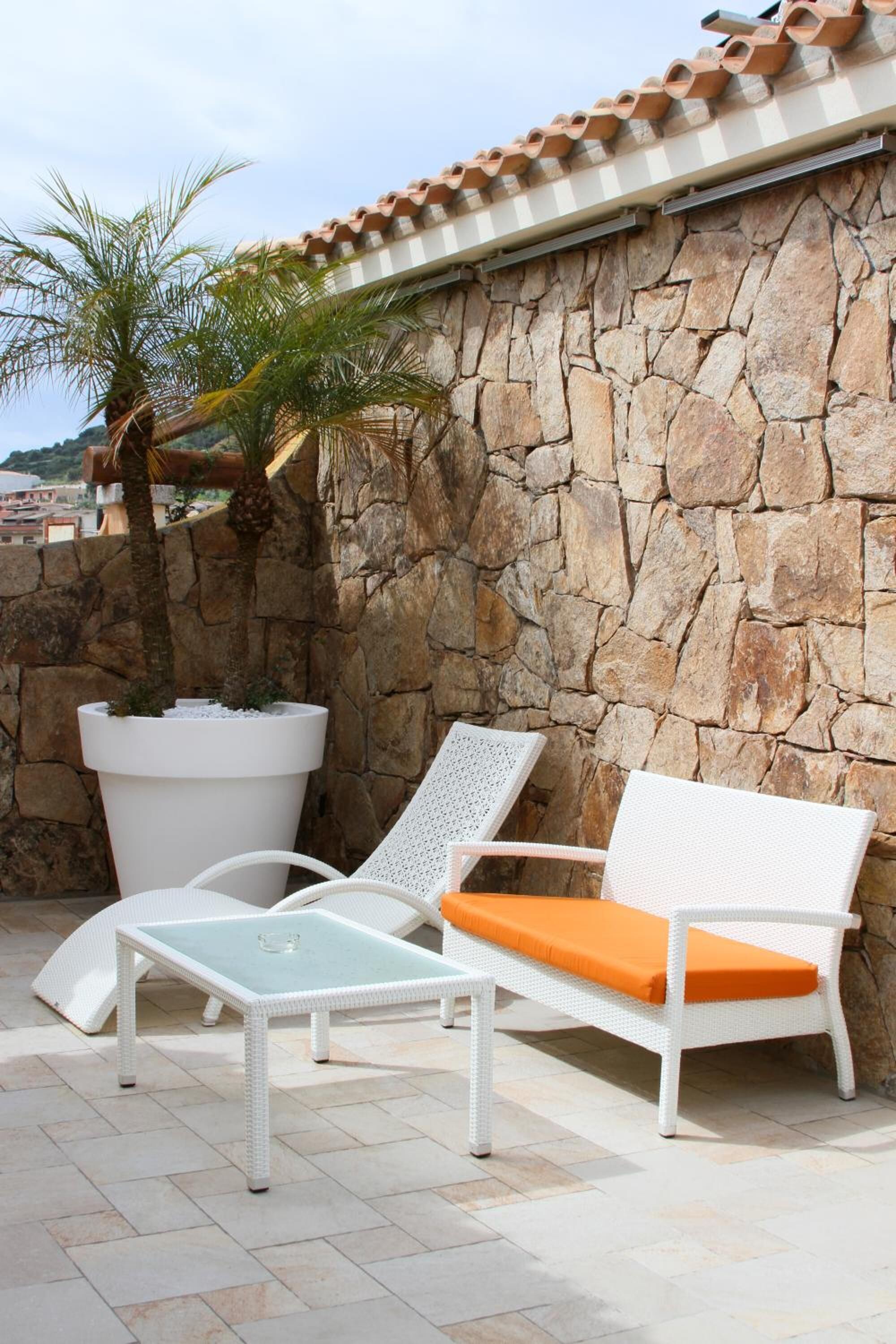 Un patio con paredes de piedra, una silla lounge de mimbre blanco con cojín naranja y una mesa con cubierta de vidrio.