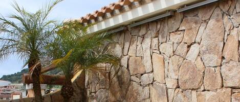 Un patio con paredes de piedra, una silla lounge de mimbre blanco con cojín naranja y una mesa con cubierta de vidrio.