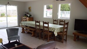 A dining area with a table set for four, wooden chairs, a bench, a television, and a window with a view of a fence and greenery.