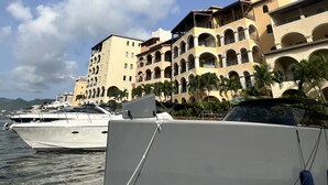 Ein Yachthafen mit mehreren dort liegenden Yachten, ein Gebäude mit mehreren Balkonen und Bögen, und ein klarer Himmel mit Wolken.