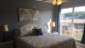 A bedroom with a large bed, two bedside tables with lamps, a painting on the wall, and a view of a dock and water through a large window. - Ledges Complex - Lake front With Slip Included - Beach and 2 Pools (Osage Beach)