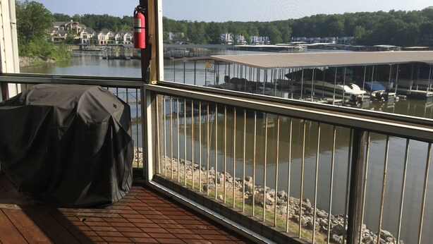 A wooden deck with a covered grill, a fire extinguisher, and a view of a marina with boats and buildings across the water. - Ledges Complex - Lake front With Slip Included - Beach and 2 Pools (Osage Beach)