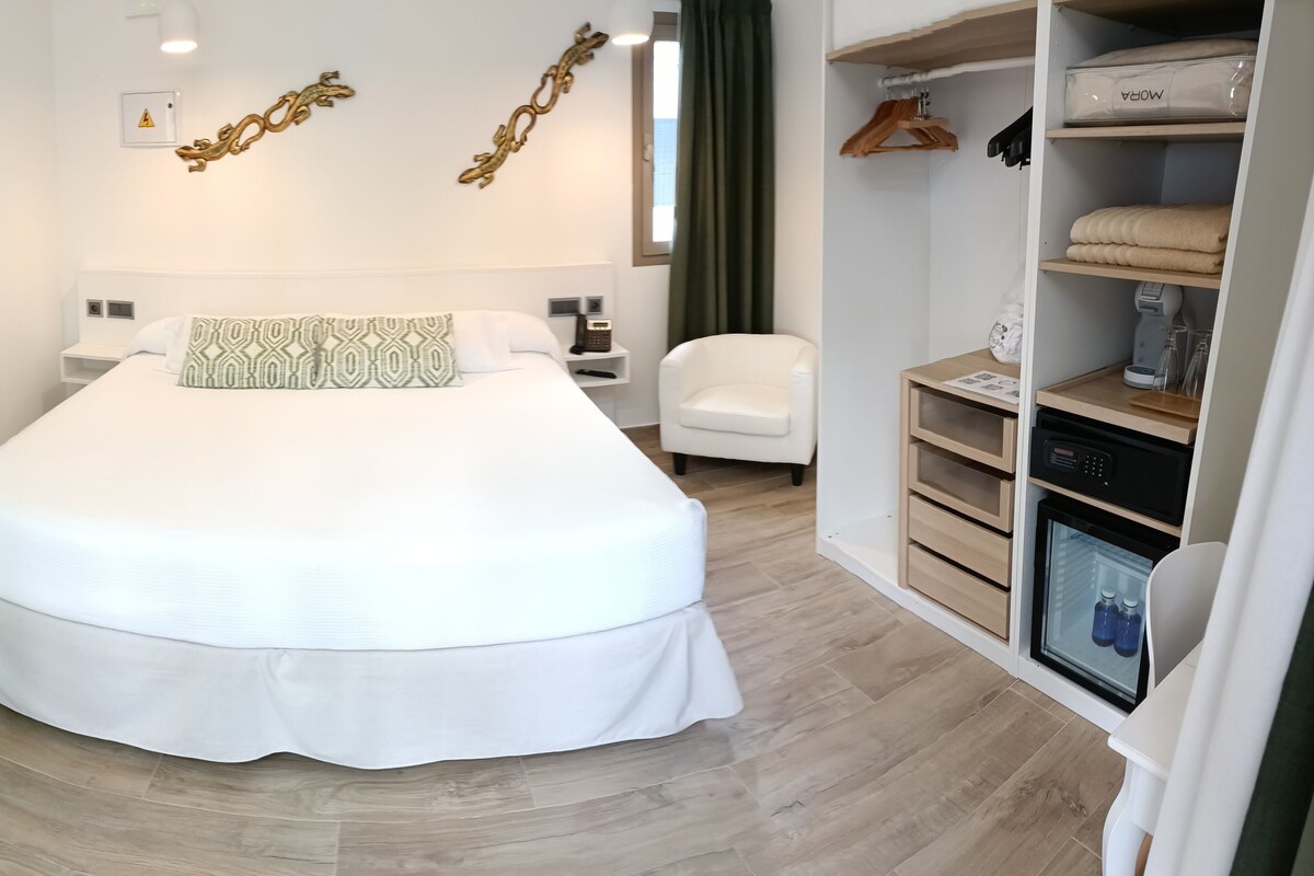 Une chambre d’hôtel moderne dotée d’un grand lit, d’une coiffeuse avec miroir, d’un petit coin salon et d’une armoire contenant un mini-bar.