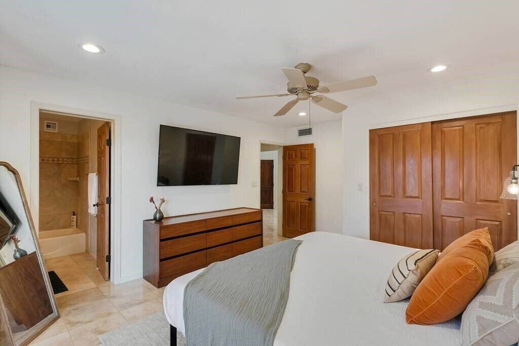 A bedroom with a bed, a dresser, a television mounted on the wall, a ceiling fan, and wooden doors.