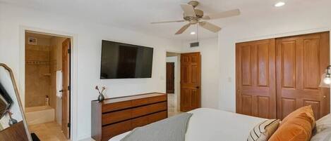 A bedroom with a bed, a dresser, a television mounted on the wall, a ceiling fan, and wooden doors.