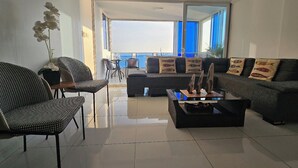 Una sala de estar moderna con sofá, sillas, una mesa de centro y vista al mar a través de amplios ventanales.