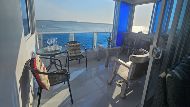 Un balcón con vista al mar, amoblado con una mesa, sillas y una pequeña planta.